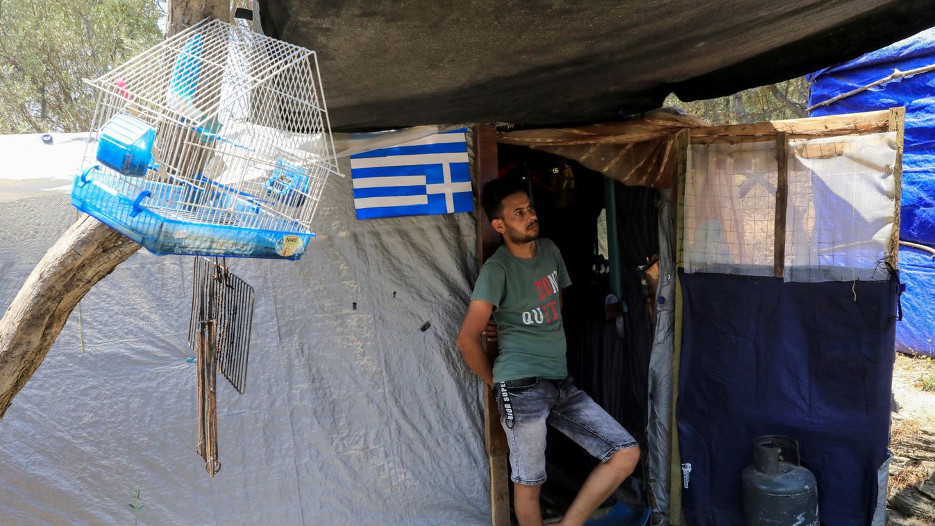 A migrant stands at the entrance of his tent at a makeshift camp for refugees and migrants next to the formal camp, over the town of Vathy on the island of Samos