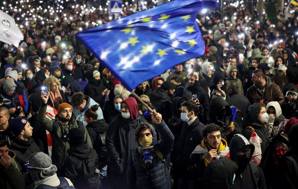  Egy tüntető az Európai Unió lobogóját lengeti, miközben tüntetők gyülekeznek a Parlament épülete mellett, ellenezve a kormány döntését, miszerint 2028-ig elhalasztja az Európai Unióhoz (EU) való csatlakozási tárgyalásokat Tbiliszi belvárosában 2024. november 29-én