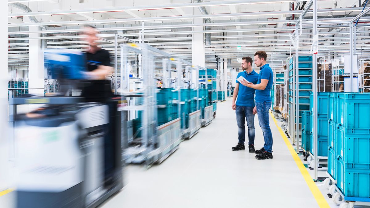 Two men with tablet talking in industrial hall while man driving a tugger train