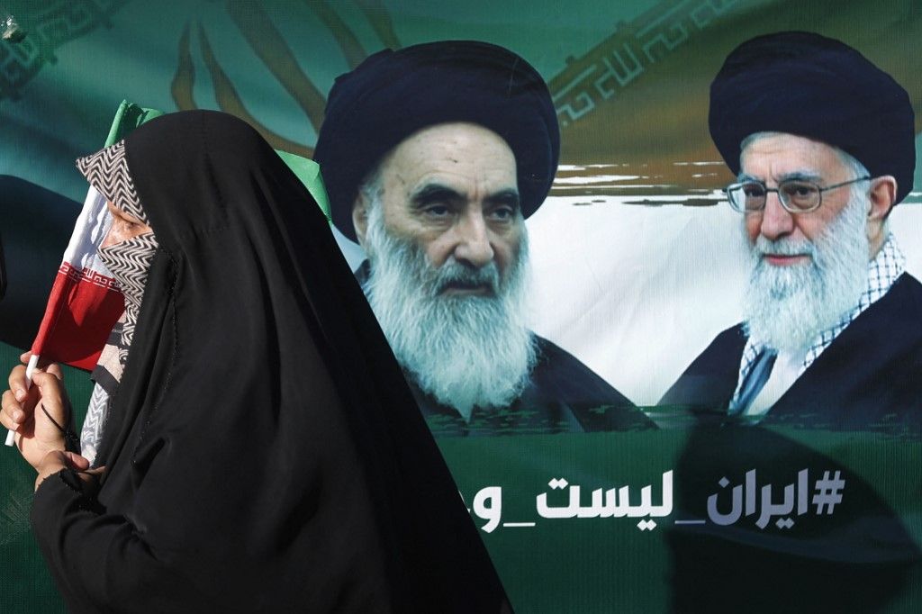 An Iraqi woman walks past a portrait of Iraq's top Shiite cleric Ayatollah Ali al-Sistani and Iran's supreme leader Ayatollah Ali Khamenei during march in the Kadhimiya district of Baghdad on June 21, 2025, to protest against Israel's strikes on Iran. (Photo by AHMAD AL-RUBAYE / AFP)