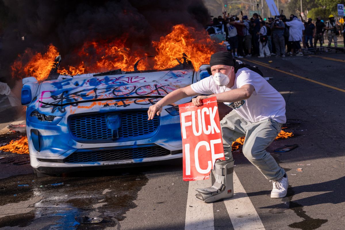 A demonstrator crouches in front of a flaming vehicle holding a sign that reads FUCK ICE in Los Angeles, California on June 8, 2025. Protests broke out across Los Angeles in response to federal immigration raids and the deployment of National Guard troops by the Trump administration. The protests, led by immigrant rights groups and local residents, quickly escalated into street clashes with law enforcement. Demonstrators waved Mexican flags, held signs against ICE, and set fire to cars in acts of defiance. (Photo by Sahab Zaribaf / Middle East Images via AFP)
