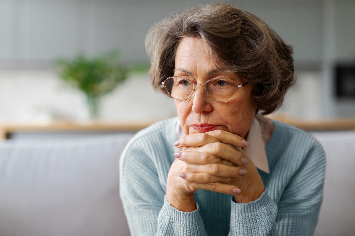 Thoughtful sad elderly woman sitting on couch, pensive older female looking into distance, thinking about problems