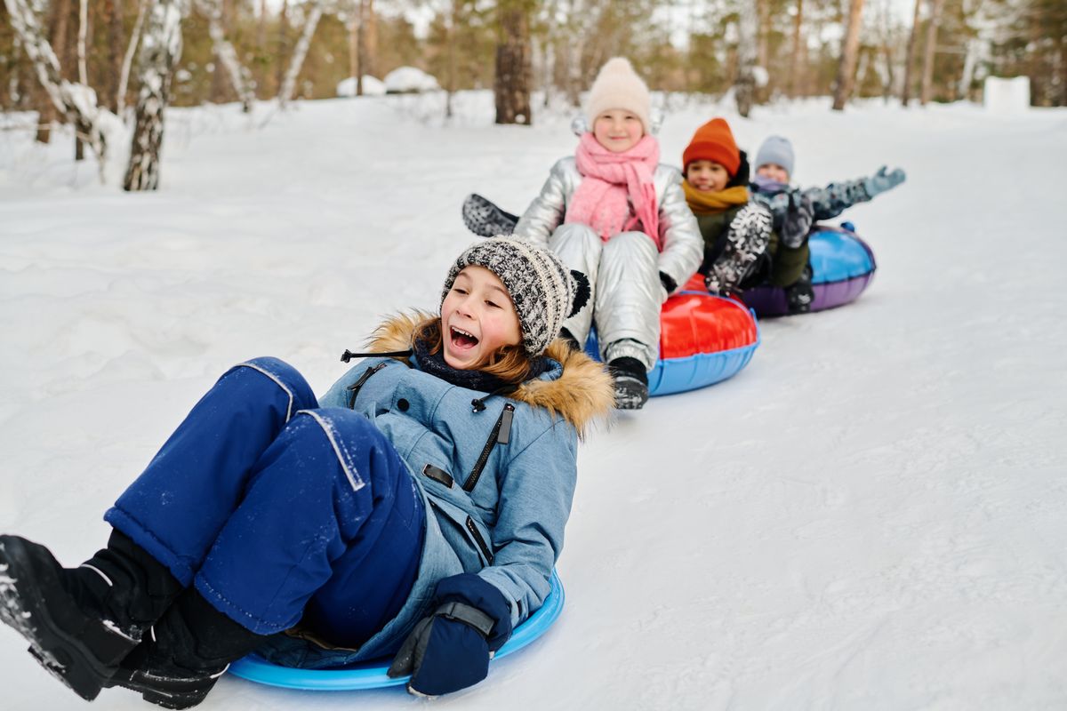 Cute excited boy in winterwear sitting on slide while moving down snow hill against group of his cheerful friends on tubes