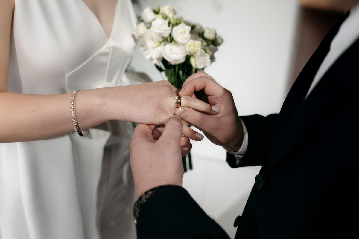 Two people exchange rings in a wedding ceremony, one placing a ring on the other's finger. Blurred flowers in the background enhance the ceremonial context.
