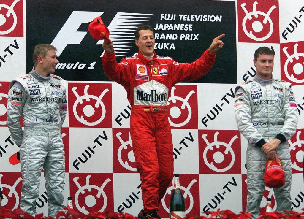 Ferrari driver Michael Schumacher (C) of Germany jokingly conducts during the playing of the Italian national anthem for Ferrari while McLaren-Mercedes driver Mika Hakkinen (L) of Finland and his teammate David Coulthard (R) of Scotland look on, during the award ceremony after Schumacher won the Formula One Japanese Grand Prix in Suzuka 08 October 2000.  Schumacher won the race to clinch the drivers' title this year, with Hakkinen finishing second and Coulthard third. AFP PHOTO/Toru YAMANAKA (Photo by TORU YAMANAKA / AFP)