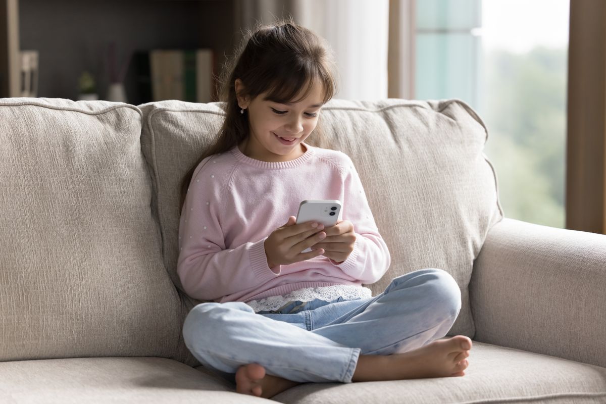 Engaged in online gaming. Smiling little child schoolgirl relax at home sitting on comfy couch using mobile phone to play games send text messages in chat surf internet under parental control software