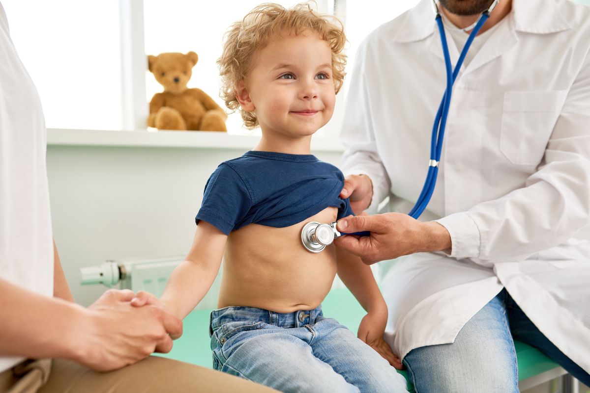 Portrait of adorable little boy visiting doctor, looking brave and smiling, holding mothers hand while pediatrician listening to heartbeat with stethoscope