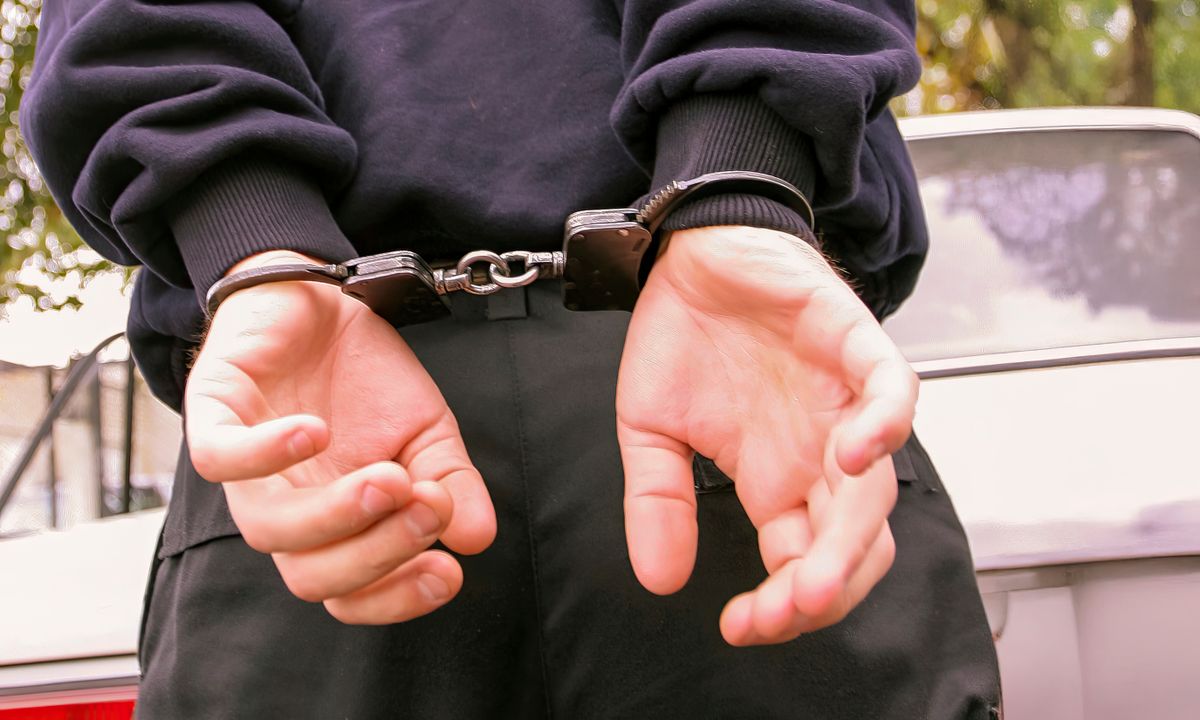 A person is in handcuffs, with hands behind their back, standing near a parked vehicle under natural lighting conditions.