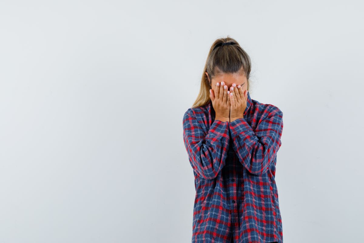 young lady covering face with hands in checked shirt and looking depressed , front view.