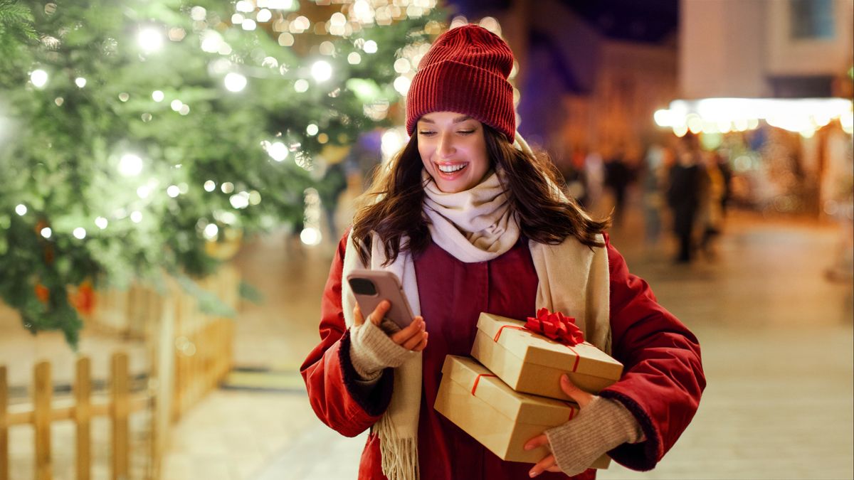 Modern holiday shopping. Happy lady holding gift boxes and using smartphone while standing outdoors near Xmas tree and festive decorations