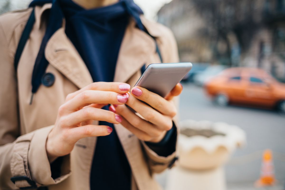 Young woman wearing beige coat using smart phone standing near road with cars in city at spring day. Close-up female's hands holding mobile phone outdoors.
