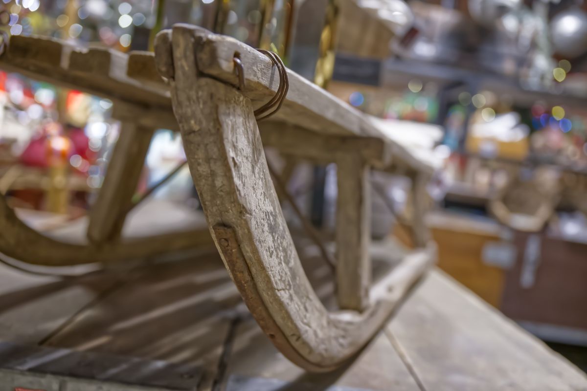 A close-up view of a vintage wooden sled, showcasing its curved runners and rustic texture. The background features a blurred assortment of antique items, creating a nostalgic atmosphere.