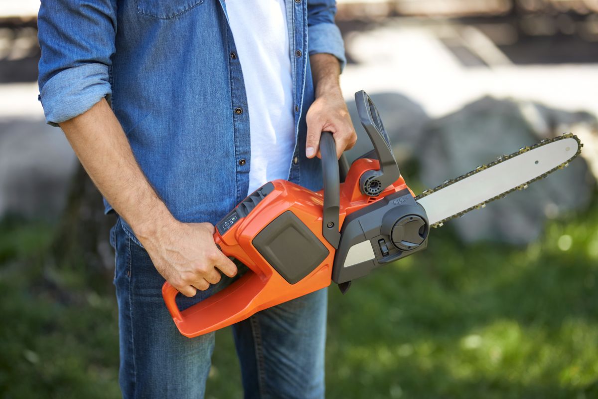Anonymous craftsman in jeans holding battery chain saw, while working in garden. Crop view of strong lumberjack using lightweight electric saw, with blurred background. Concept of pro equipment.