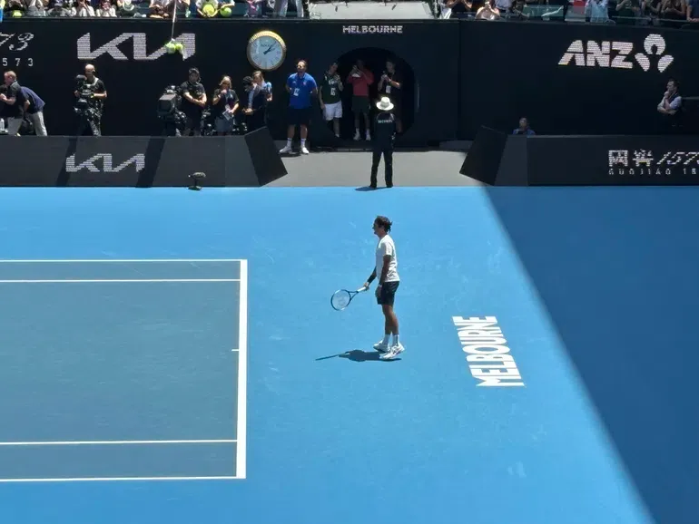 Roger Federer az alapvonal mögül várja a labdát, a kijáratból Novak Djokovics figyeli az edzést az Australian Openen