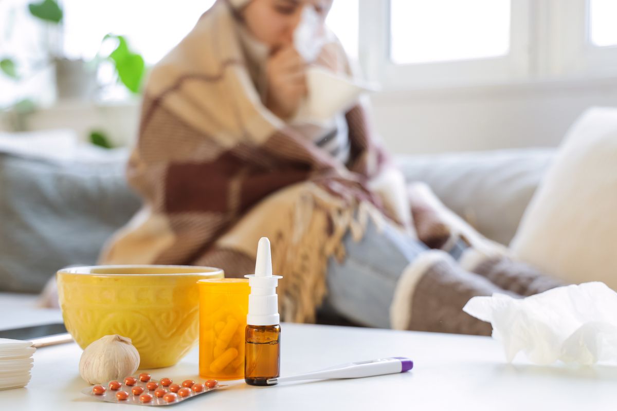 Different flu medications with garlic on table against sick woman at home, closeup