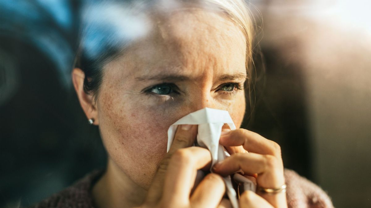 Ill woman sneezing into tissue paper while standing behind a window.