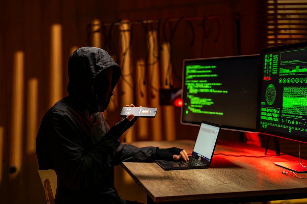 Cybercriminal in a dark hooded sweatshirt working on a laptop and holding a smartphone. With multiple monitors displaying code and graphs. Representing cybersecurity threats and data privacy concerns
