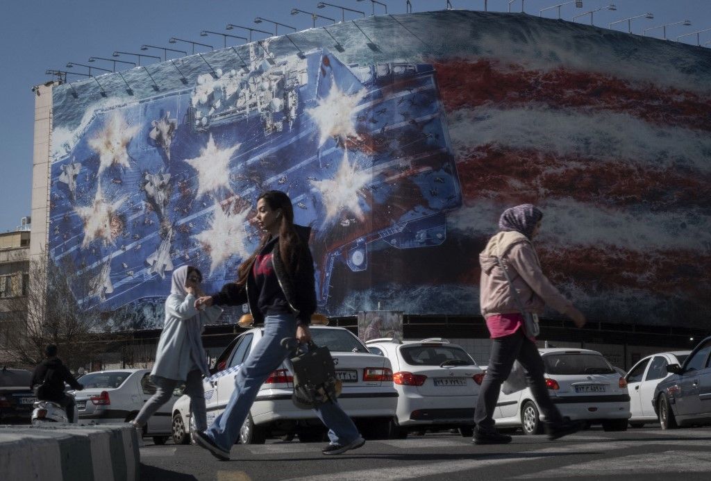 Fiatal iráni nők sétálnak el egy állami épület előtt, amelyet egy hatalmas, az Egyesült Államokat ellenző óriásplakát borít, amelyen a megsemmisült USS Abraham Lincoln (CVN-72) repülőgép-hordozó szimbolikus képe látható Teherán belvárosában, Iránban