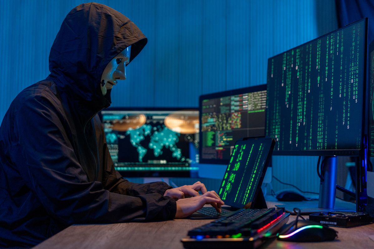 Anonymous computer hacker in white mask and hoodie. Obscured dark face using laptop computer for cyber attack and calling on cellphone, Data thief, internet attack, darknet and cyber security concept.