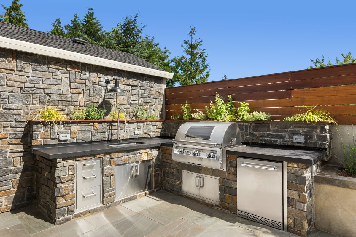 Modern outdoor kitchen area featuring a built in stainless steel gas grill with a black countertop and stone veneer walls under a clear blue sky