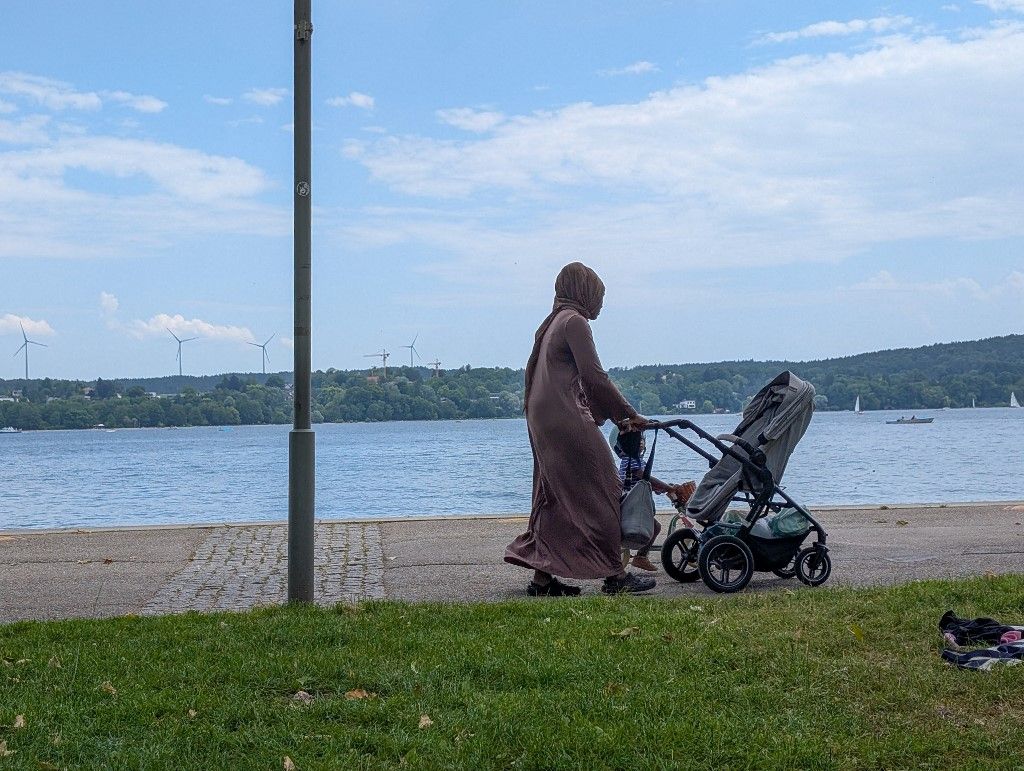A woman in a long dress and headscarf walks with a young child and pushes a stroller along the shore promenade of Lake Starnberg in Starnberg, Bavaria, Upper Bavaria, Germany, on June 15, 2025. Wind turbines and sailboats are visible across the lake. (Photo by Michael Nguyen/NurPhoto) (Photo by Michael Nguyen / NurPhoto via AFP)