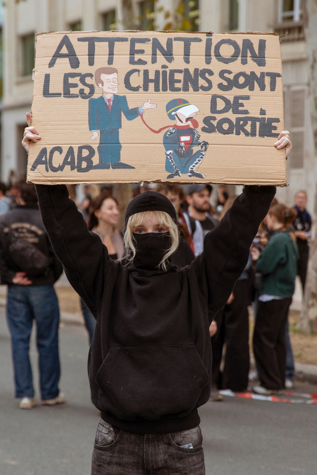 A masked protester raises a cardboard sign reading "Beware, the dogs are out" above their head in Paris, France, on October 13, 2026. The demonstration was part of nationwide student-led protests demanding more funding and better conditions in education, during which police used tear gas and water cannon near Les Invalides. (Photo by Davood Maeili / Middle East Images via AFP)